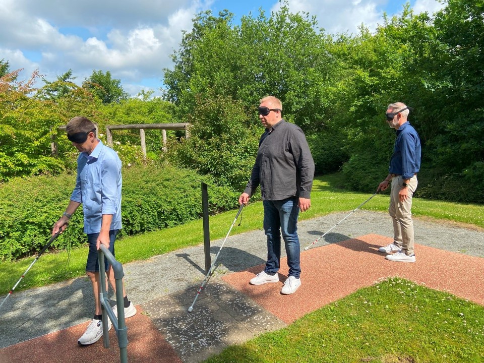 2 mandlige synskonsulenter og en mandlig optometrist har sorte bind for øjnene og går gennem en forhindringsbane med mobilitystokke i hænderne. De virker koncentrerede og rolige.