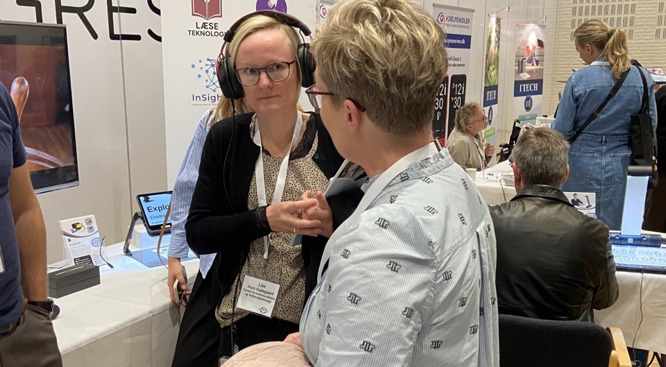 To kvindelige synskonsulenter afprøver lyd fra læseskanner på Synsmessen. De ser koncentreret på hinanden, mens den ene lytter med høretelefoner på.