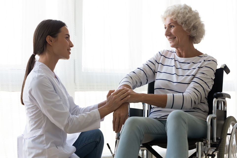 Ung kvinde i kittel knæler foran ældre kvinde i kørestol og holder hendes hånd